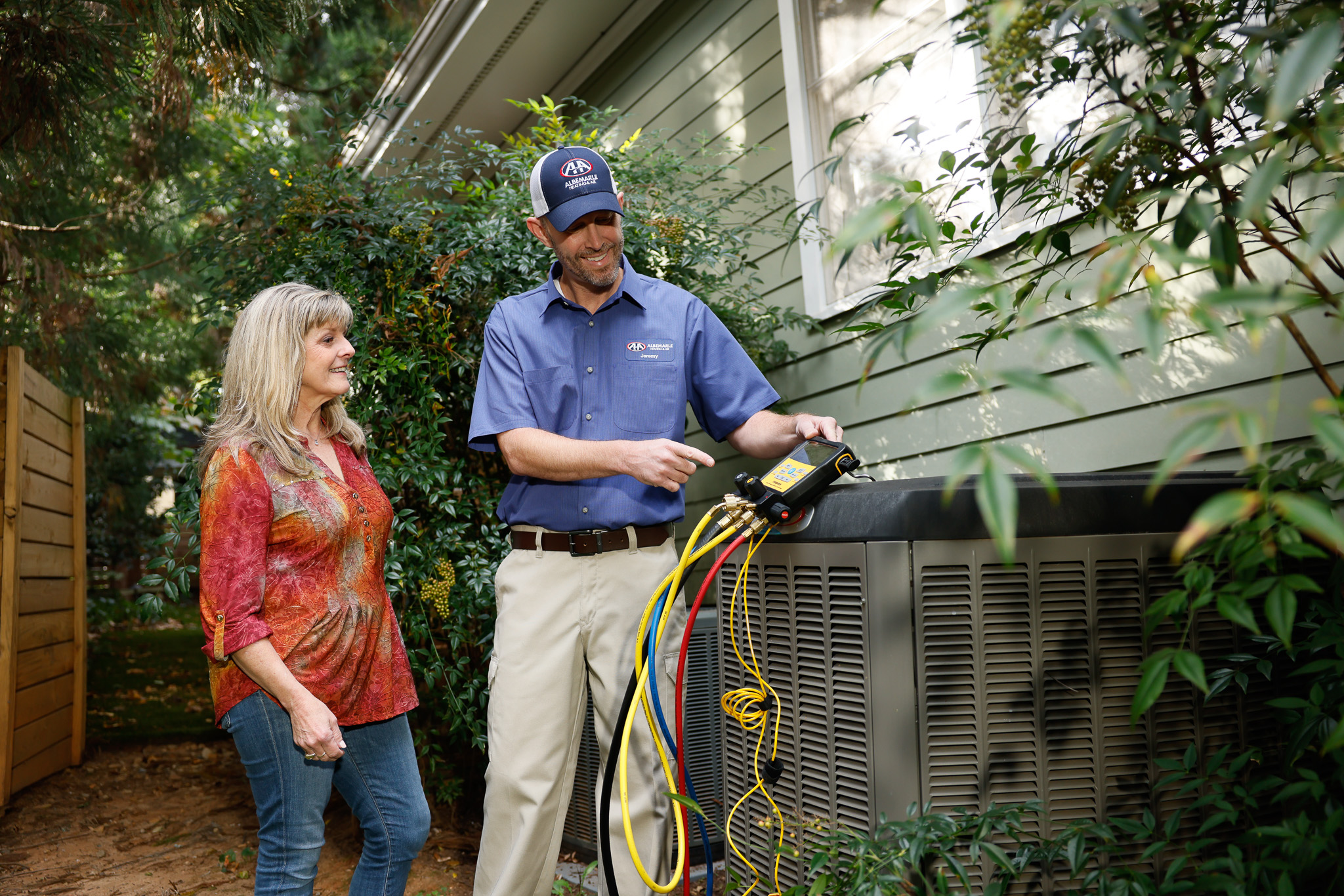 Technician speaking with a homeowner while showing her the gauges attached to the outdoor HVAC unit, next to a home with green siding.